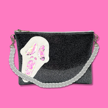 Glitter infused black vinyl bag with a white 'Ghostface' applique with pink confetti weed leaves for the eyes, nose, and mouth. Clear acrylic chain crossbody strap is connected to the top of both ends of the bag. Bag is pictured on a bright pink background.