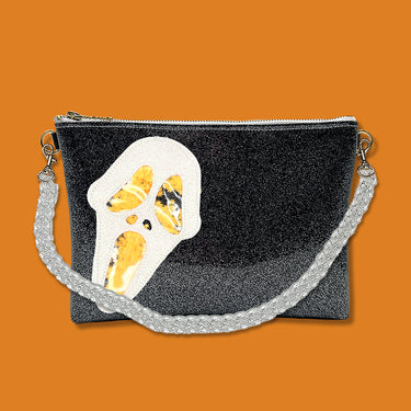 Glitter infused black vinyl bag with a white 'Ghostface' applique with orange confetti weed leaves for the eyes, nose, and mouth. Clear acrylic chain crossbody strap is connected to the top of both ends of the bag. Bag is pictured on a bright orange background.