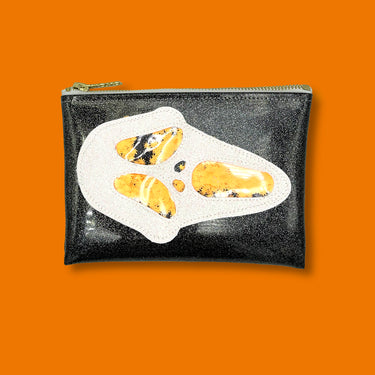 Glitter infused black vinyl clutch with a white 'Ghostface' applique with orange confetti weed leaves for the eyes, nose, and mouth. Clutch is adorned on top with a cute XOXO zipper pull that is a signature of the Julie Mollo! brand. Clutch is featured on a bright orange background. 