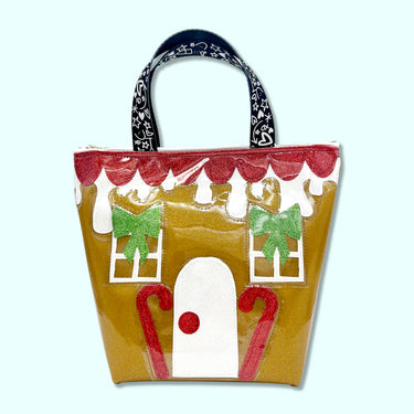 Super cute gingerbread handle bag featuring vibrant green, white, and red glitter canvas details, this charming gingerbread house sparkles on a rich gingerbread-gold glitter vinyl bag. Finished with Julie Mollo! doodle-printed handles, this unique accessory is the perfect signature winter holiday statement piece. Bag is pictured on an off-white background.