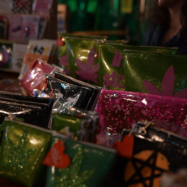 Julie Mollo posing with a collection of kush kard x Julie Mollo clutches, makeup bags, and cosmetic bags. Vibe of photo is vibey and low lit. 