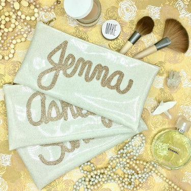 Various customized glitter infused clutches with a champagne gold name applique. Makeup tools scattered around image.